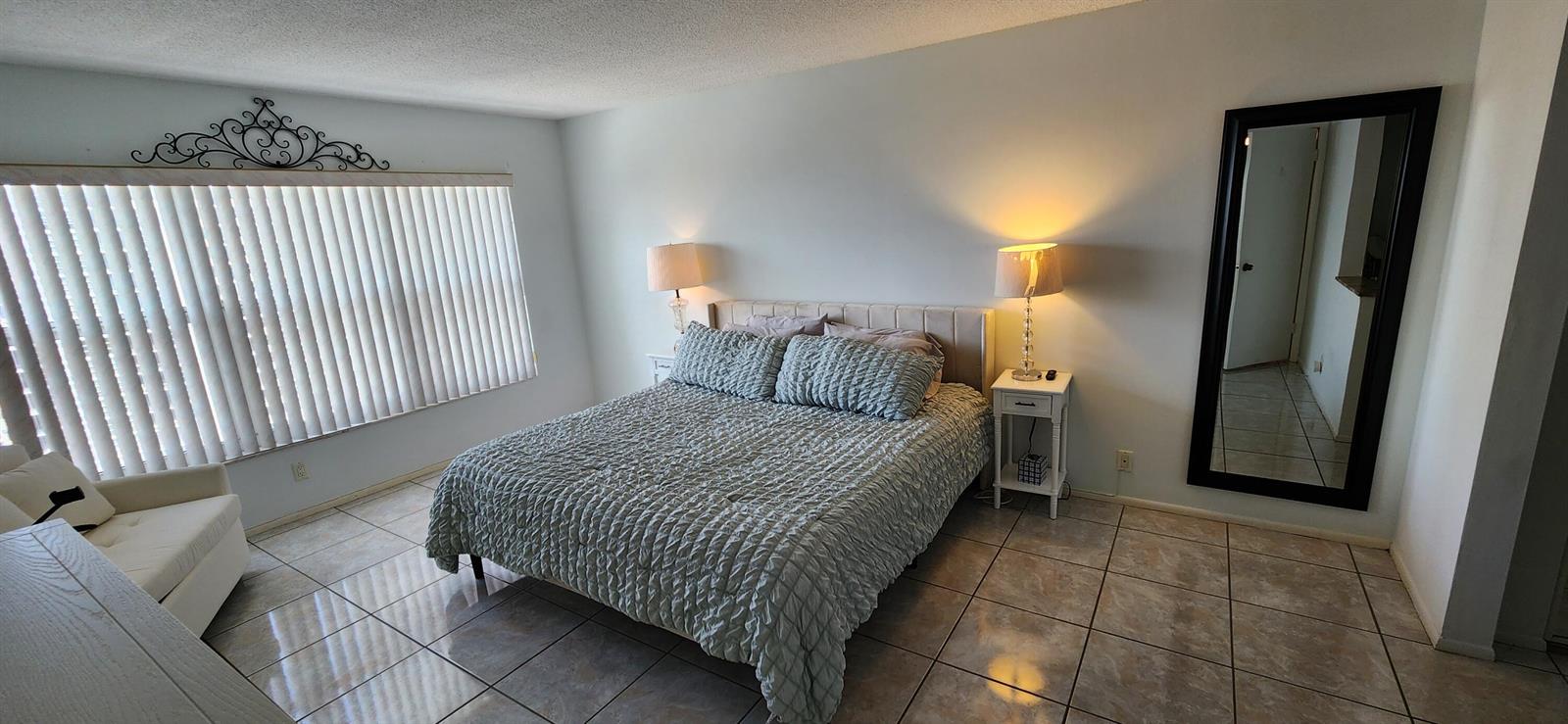 A bedroom features a bed with a textured blue comforter, tile flooring, and a floor mirror — 405 N Ocean Boulevard # 1507, Pompano Beach FL 33062, POMPANO BEACH, FL