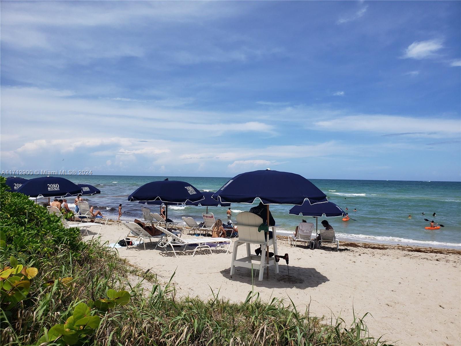 A sandy beach features rows of blue umbrellas, lounge chairs, and a lifeguard chair — 2080 S Ocean Dr # 311, Hallandale Beach FL 33009, HALLANDALE BEACH, FL