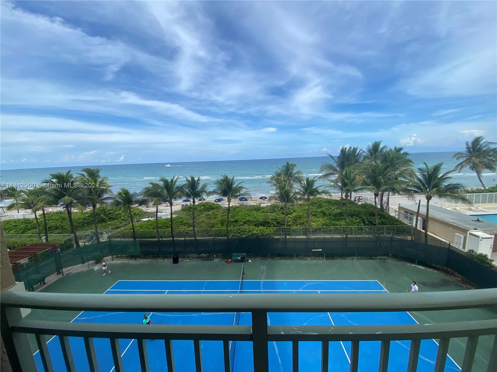 A balcony overlooks a blue tennis court with the ocean and palm trees in the background — 2080 S Ocean Dr # 311, Hallandale Beach FL 33009, HALLANDALE BEACH, FL