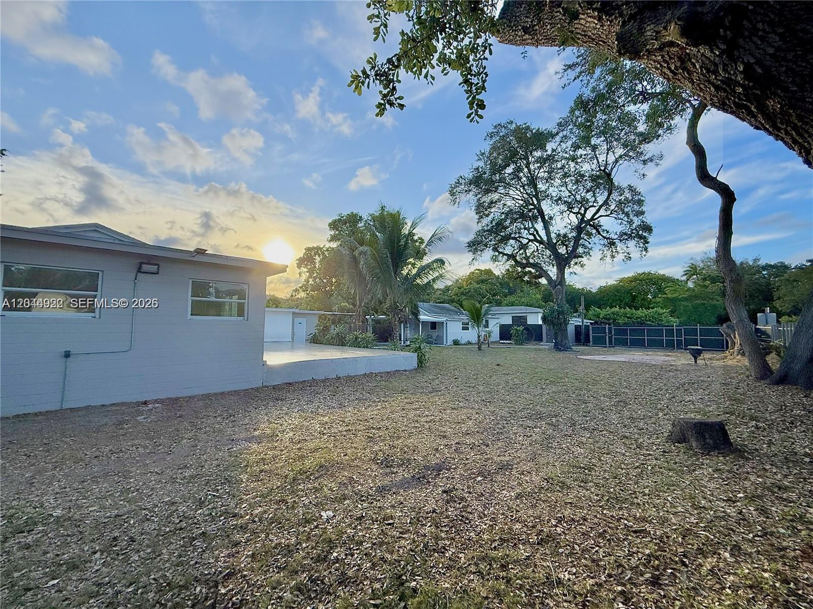 A large backyard features a light-colored house, a patio, mature trees, and a fence — 947 NE 149th St, Miami FL 33161, MIAMI, FL