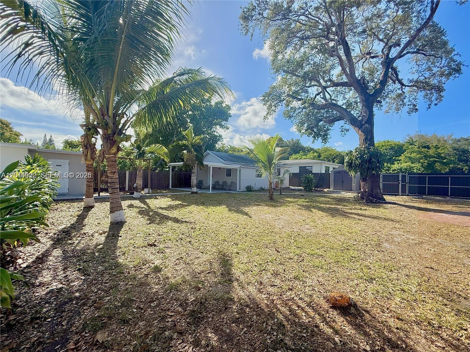 A backyard features a grassy lawn, several palm trees, a large oak tree, and houses — 947 NE 149th St, Miami FL 33161, MIAMI, FL