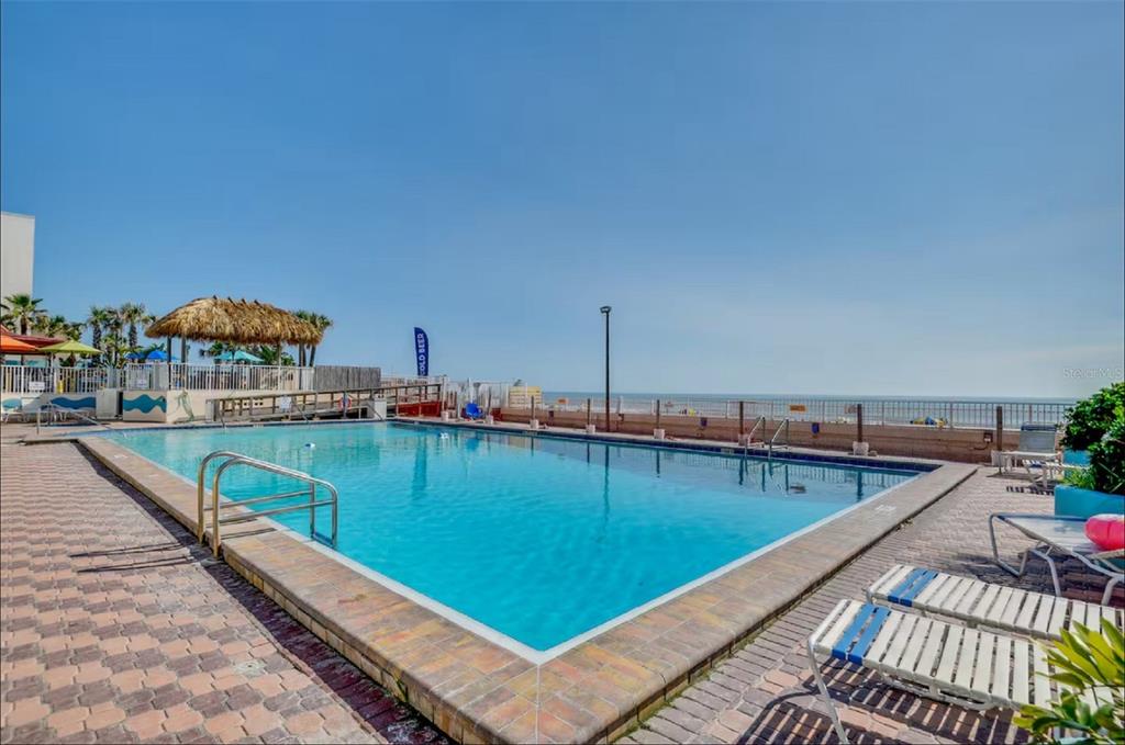 A rectangular swimming pool sits on a brick patio overlooking the ocean under a clear sky — 313 S ATLANTIC AVENUE UNIT 4100, DAYTONA BEACH, FL