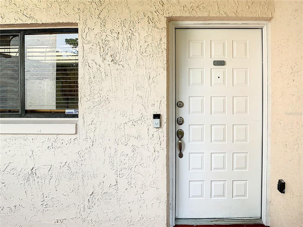 A white front door with a doorbell camera sits next to a window on a stucco wall — 1158 CARMEL CIRCLE UNIT 430, CASSELBERRY, FL