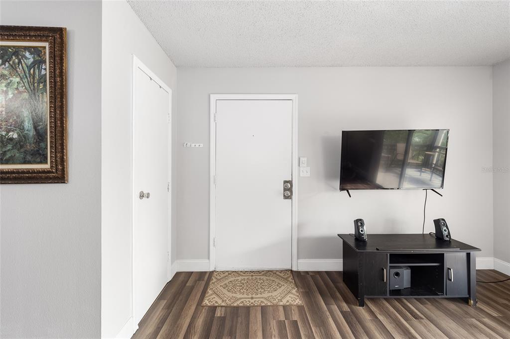 A living room features a white front door, a wall-mounted television, and wood-look flooring — 6519 W NEWBERRY ROAD UNIT 1210, GAINESVILLE, FL