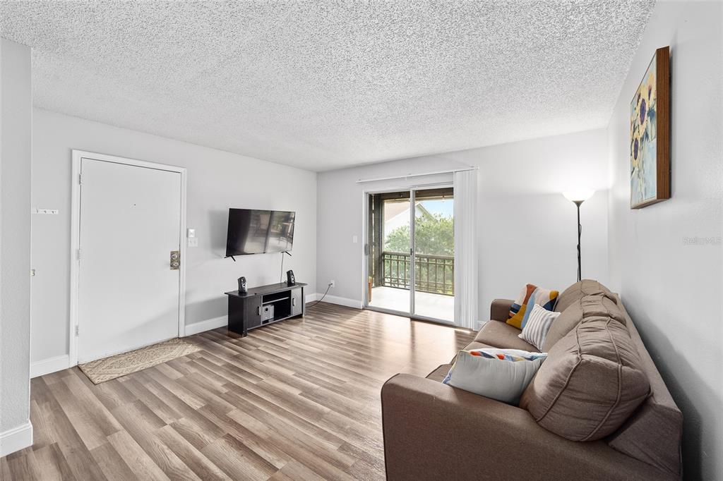 A living room features wood-look flooring, a brown sofa, a wall-mounted television, and sliding doors — 6519 W NEWBERRY ROAD UNIT 1210, GAINESVILLE, FL