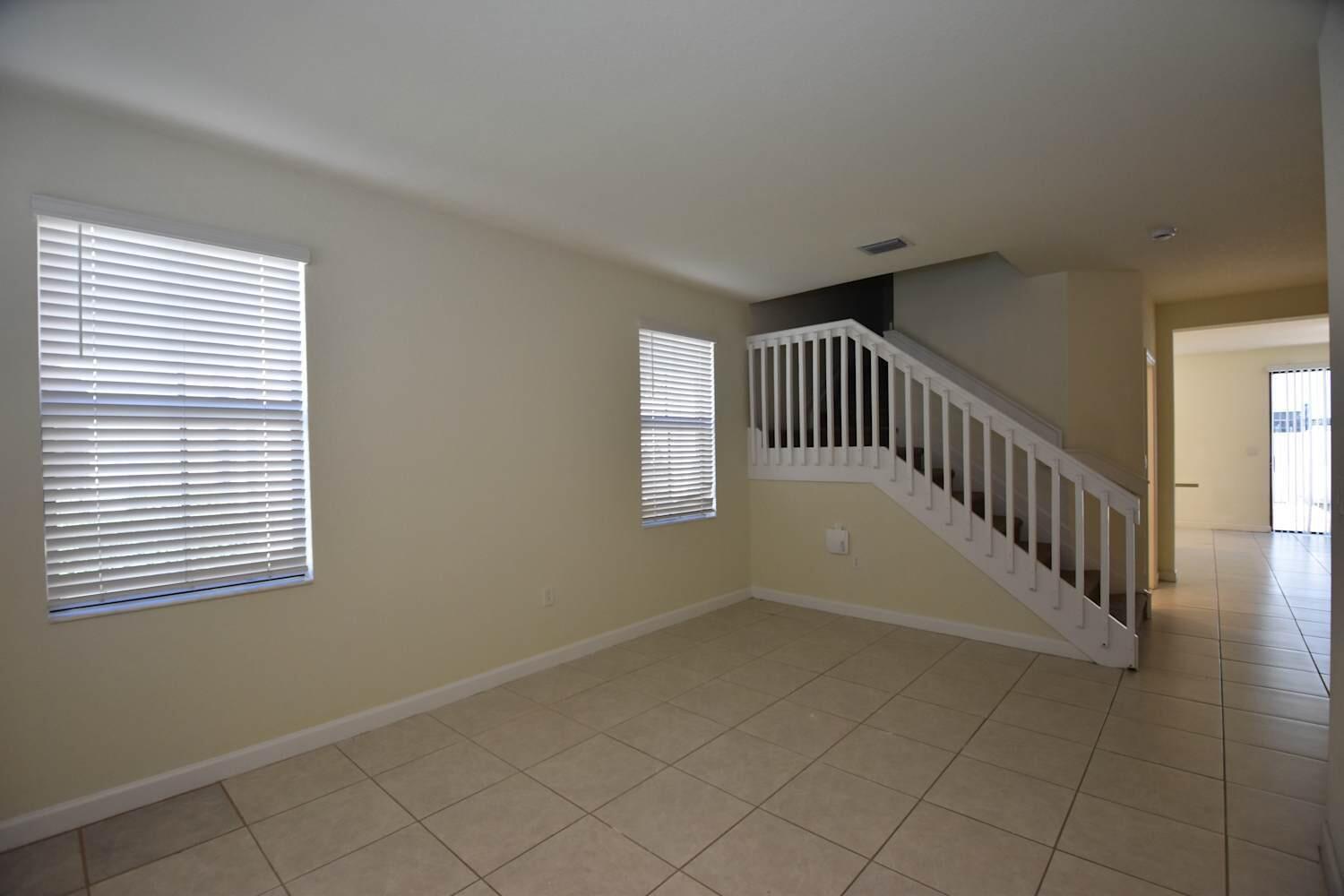 An empty living room features beige tile flooring, two windows with blinds, and a staircase — HIALEAH, FL