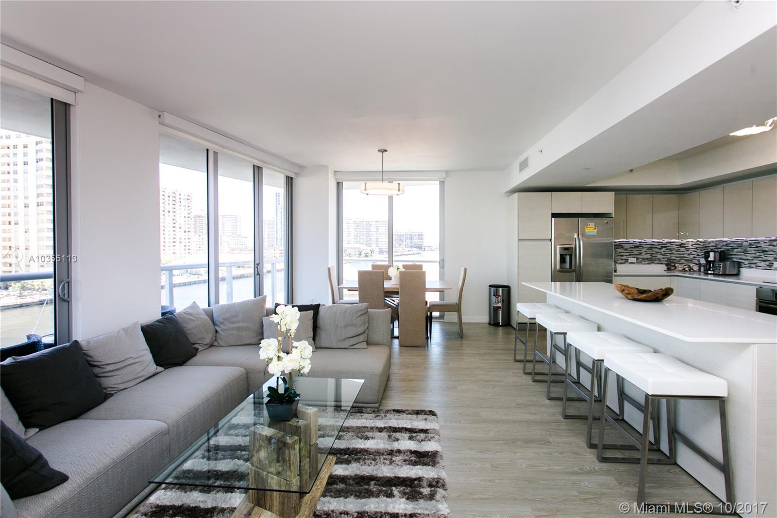 A modern open-concept living and dining area features a gray sectional, kitchen island, and balcony — 2600 E Hallandale Beach Blvd, Hallandale Beach FL 33009, HALLANDALE BEACH, FL