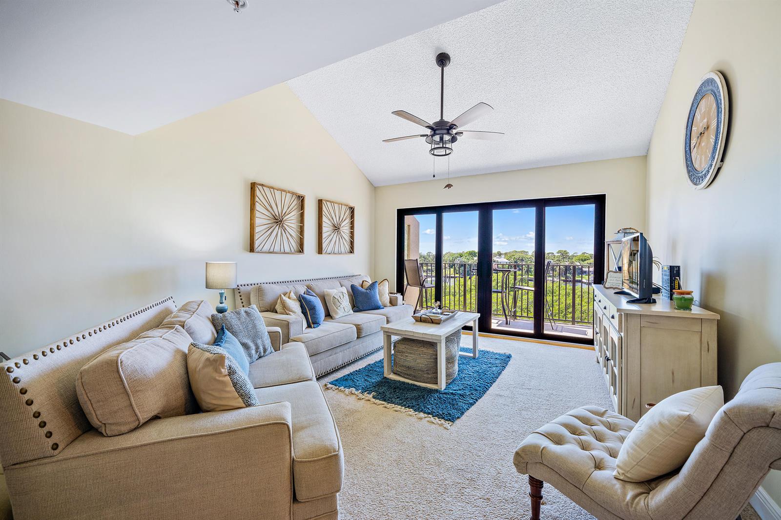 A living room features a vaulted ceiling, beige sofas, a blue rug, and sliding glass doors — 1701 Marina Isle Way # 504, Jupiter FL 33477, JUPITER, FL