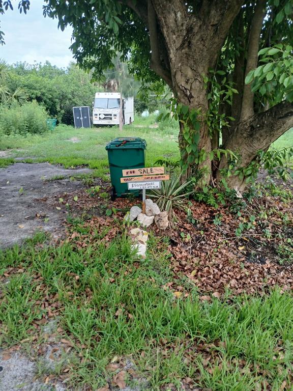 A green trash bin sits next to a for sale sign under a large tree — 5498 WILSON DRIVE, PUNTA GORDA, FL