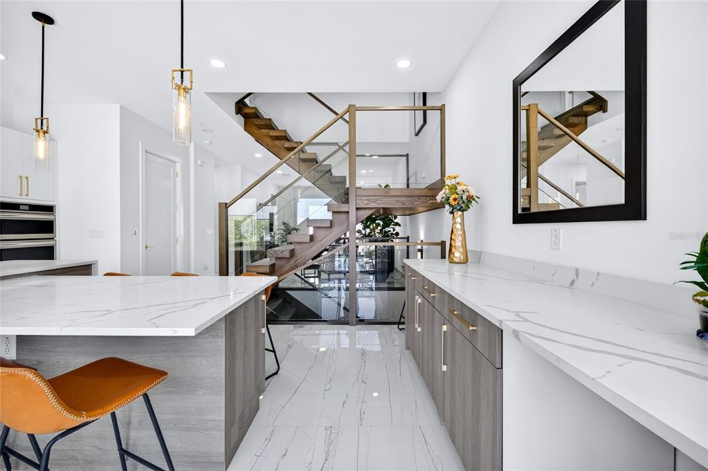 A modern kitchen features a marble-topped island, wood cabinetry, and a glass-railed staircase — 2187 MAIN STREET, SARASOTA, FL