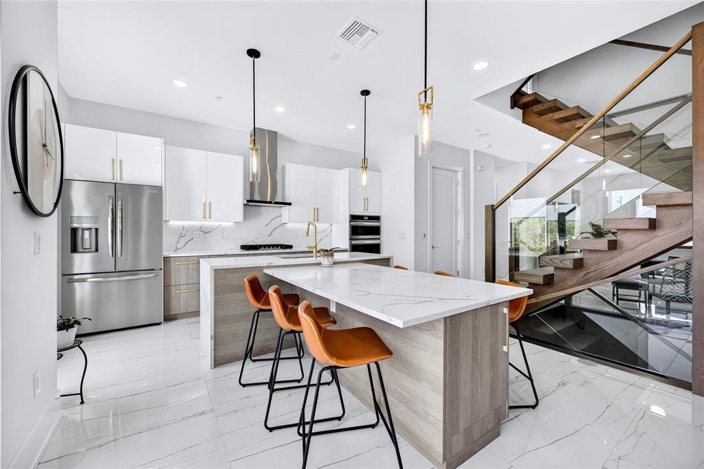A modern kitchen features white cabinetry, a marble-topped island with orange stools, and wooden stairs — 2187 MAIN STREET, SARASOTA, FL