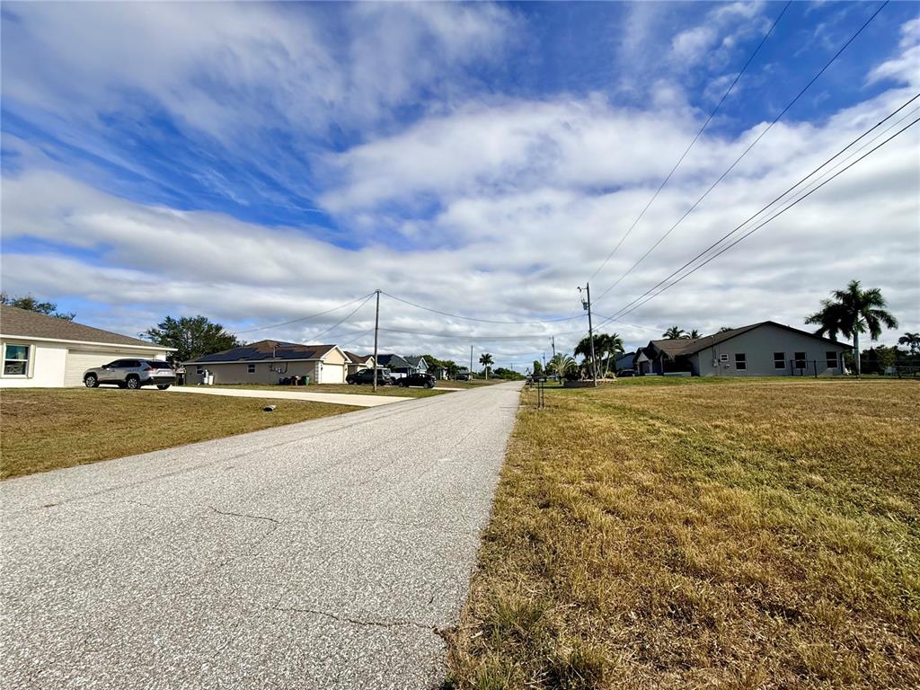 A paved residential street stretches between houses and open grassy lots under a cloudy sky — 418 NW 14TH TERRACE, CAPE CORAL, FL