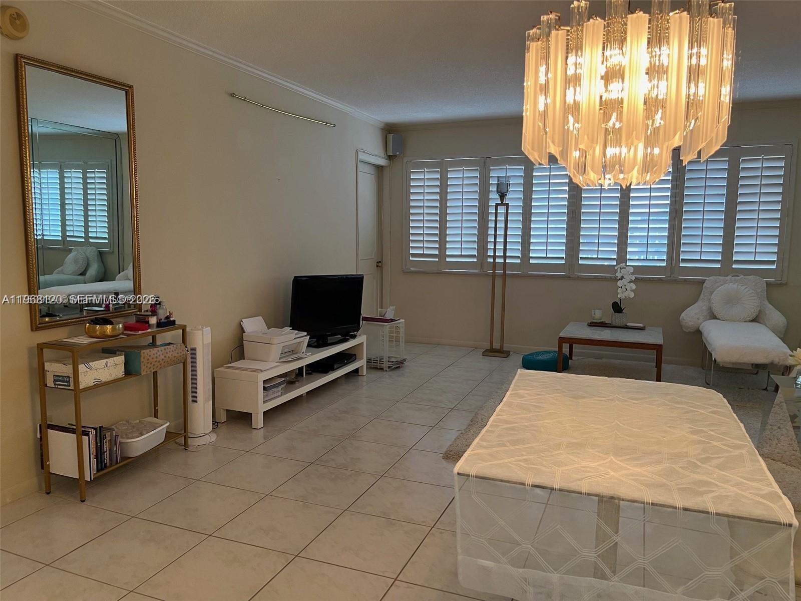 A living room features tile flooring, a large chandelier, a television, and window shutters — 215 SE 3rd Ave # D, Hallandale Beach FL 33009, HALLANDALE BEACH, FL