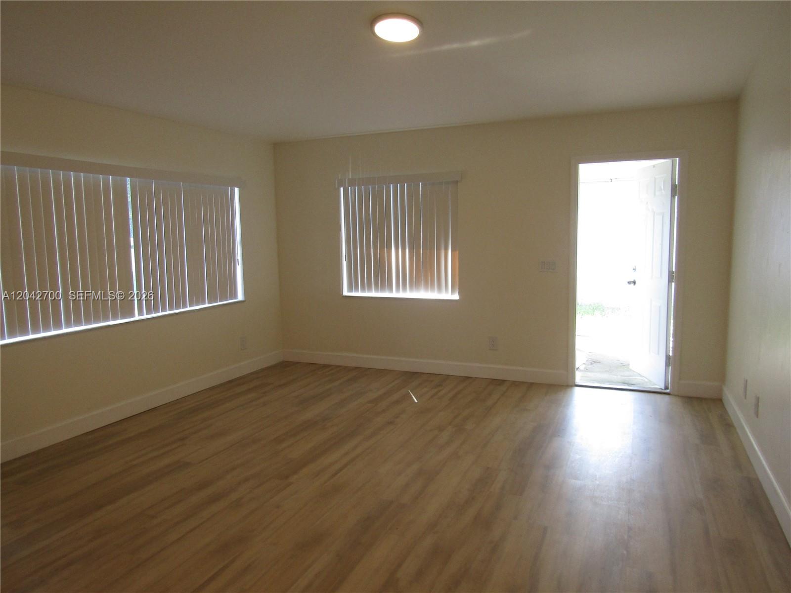 An empty room features light-colored walls, wood-look flooring, vertical blinds on windows, and an open door — 4525 28 ST SW # 4525, Lehigh Acres FL 33973, LEHIGH ACRES, FL