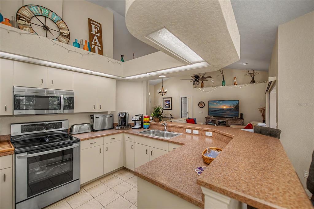 A kitchen with light-colored cabinets, stainless steel appliances, and a peninsula countertop overlooks a living area — 113 & 121 RED CEDAR PARK, ROTONDA WEST, FL