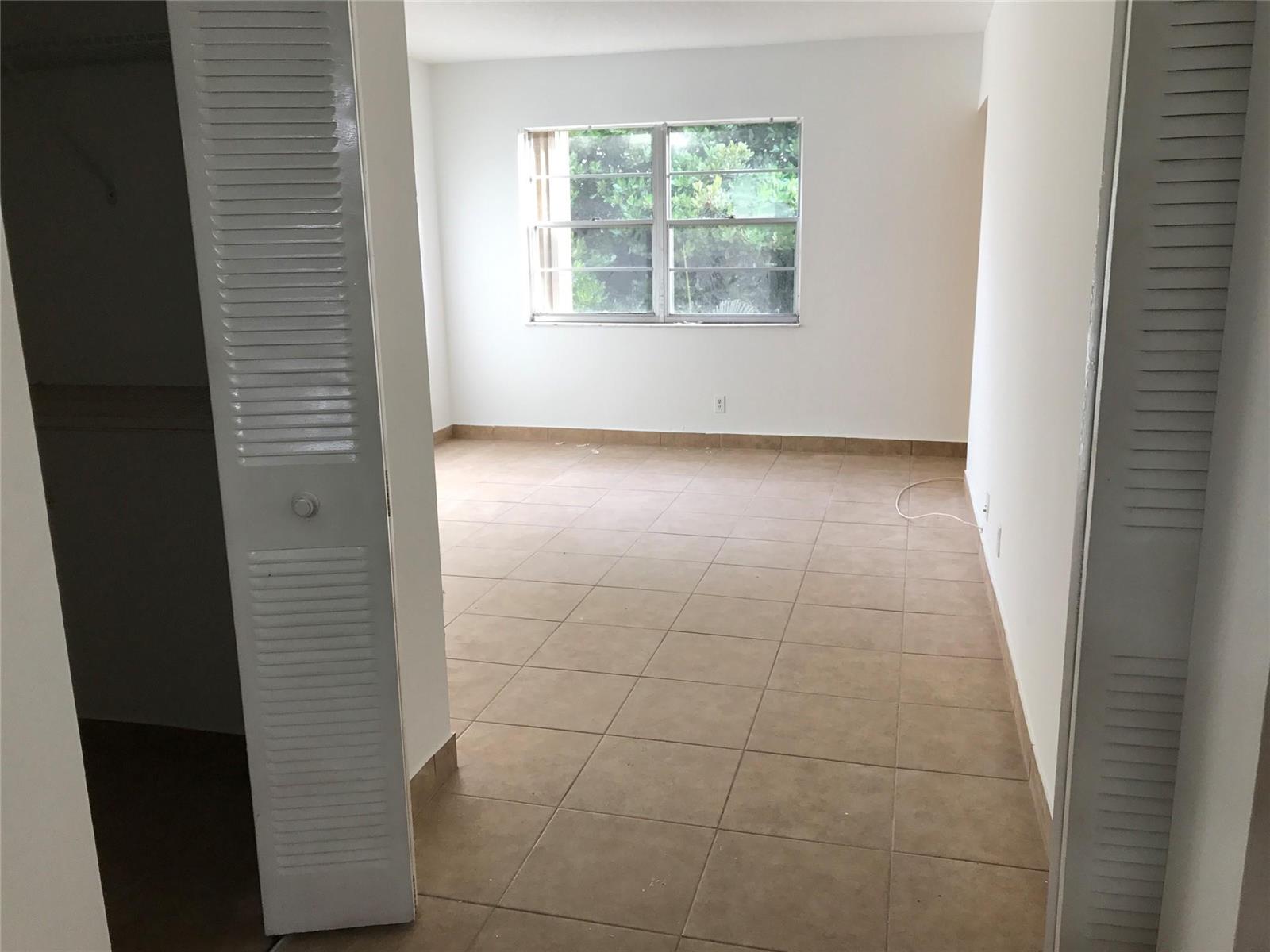 An empty bedroom features white walls, tan tile flooring, and a window overlooking greenery — 4394 NW 9th Avenue # 3a, Deerfield Beach FL 33064, DEERFIELD BEACH, FL