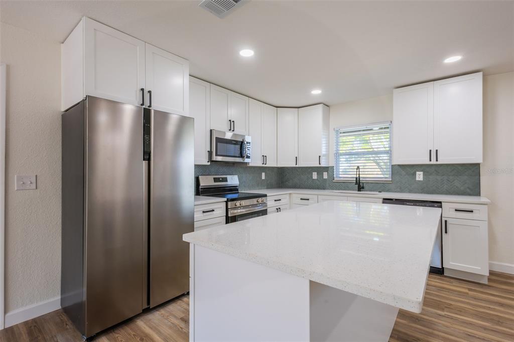 A kitchen features white cabinets, stainless steel appliances, a quartz island, and a herringbone backsplash — 1517 HALLAM COURT N, LAKELAND, FL
