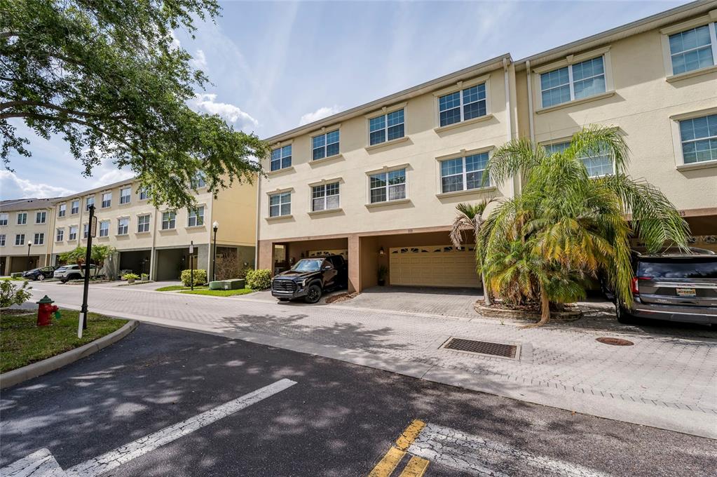 A three-story townhouse exterior features a ground-level garage, beige stucco walls, and paved parking — 10144 ARBOR RUN DRIVE UNIT 112, TAMPA, FL