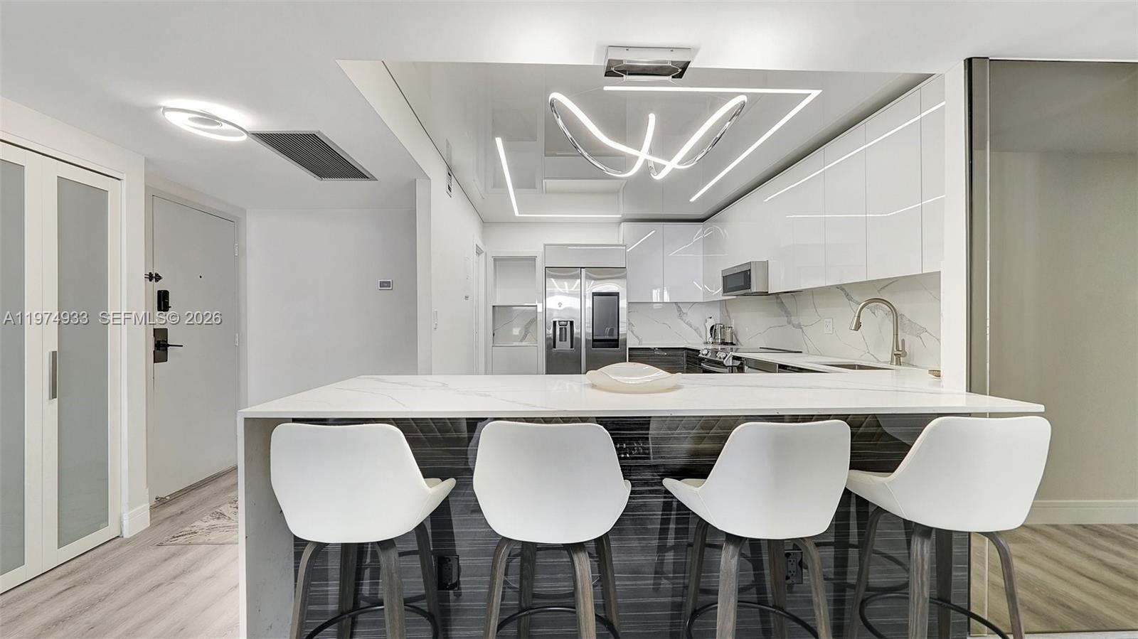 A modern kitchen features white cabinetry, a marble-topped island, and four white bar stools — 2500 Parkview Dr # 1914, Hallandale Beach FL 33009, HALLANDALE BEACH, FL