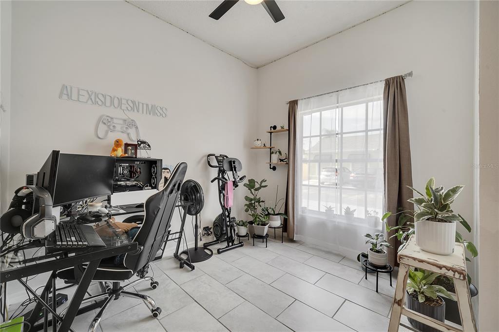 A home office with a gaming setup, exercise bike, and several potted plants on tile flooring — 1028 UNIVERSAL REST PLACE, KISSIMMEE, FL