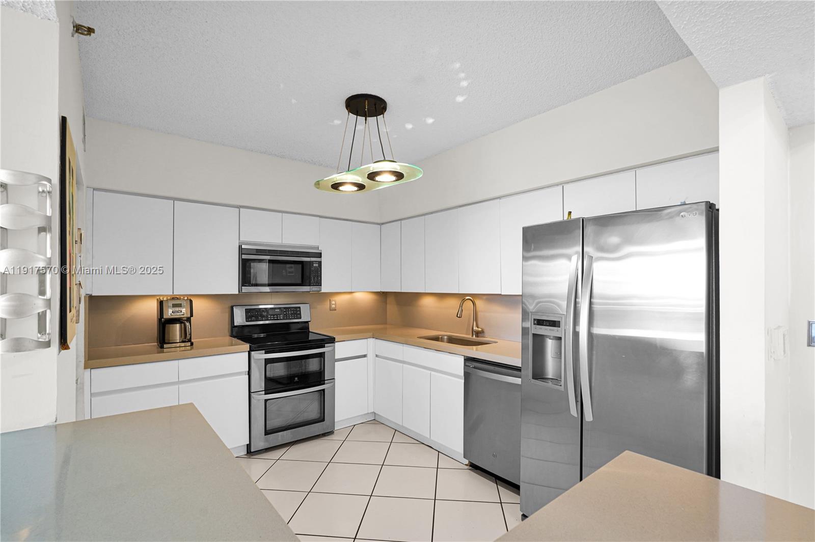 A kitchen features white cabinets, stainless steel appliances, tan countertops, and a modern light fixture — 9455 Collins Ave # 603, Surfside FL 33154, SURFSIDE, FL