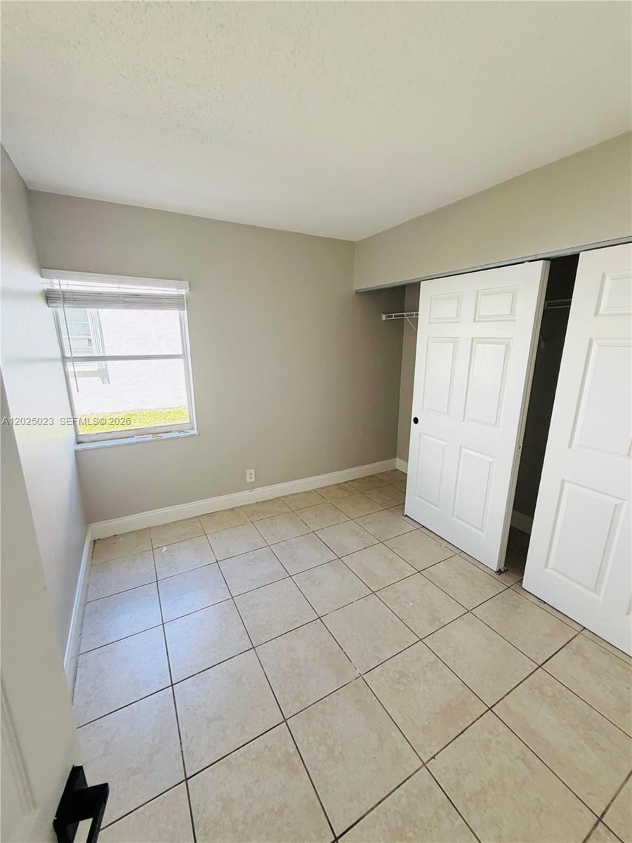 A small bedroom features light-colored walls, beige tile flooring, a window, and a closet — 331 NW 42ND CT # 101, Pompano Beach FL 33064, POMPANO BEACH, FL