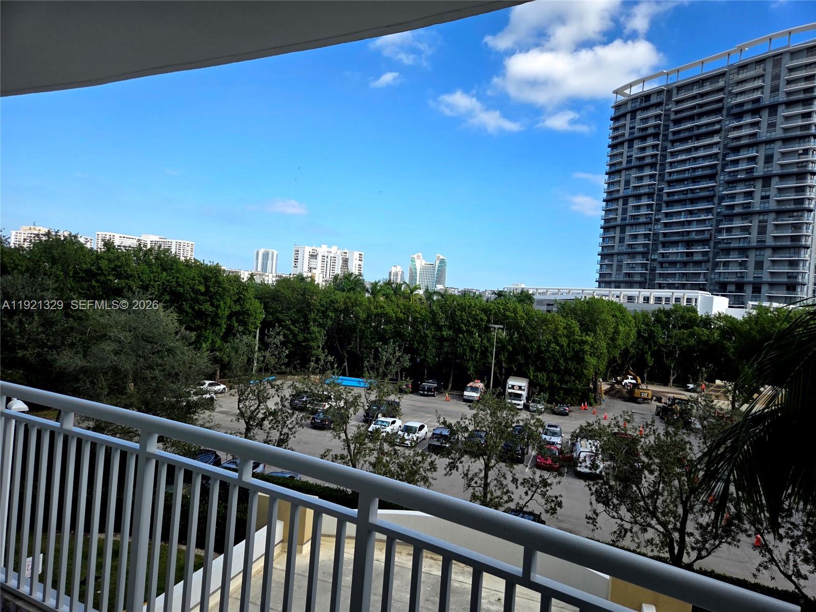 A balcony overlooks a parking lot, trees, and distant high-rise buildings under a blue sky — 1755 E Hallandale Beach Blvd # 305E, Hallandale Beach FL 33009, HALLANDALE BEACH, FL
