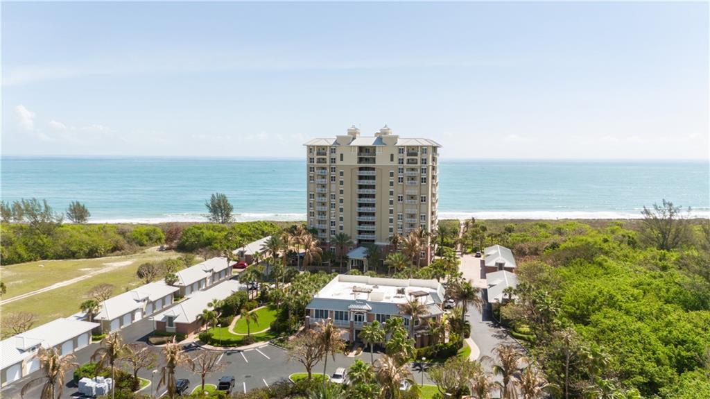 An aerial view shows a tall beachfront condominium building surrounded by lush vegetation and parking — HUTCHINSON ISLAND, FL