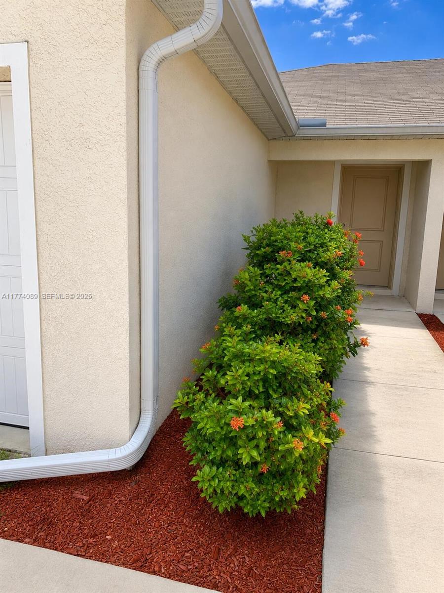 A beige stucco exterior wall features a white downspout and a flowering shrub in mulch — 9084 Aegean Cir, Lehigh Acres FL 33936, LEHIGH ACRES, FL