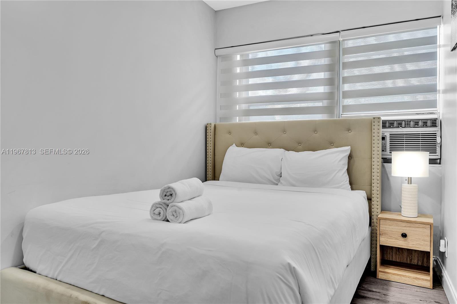 A bedroom features a tufted beige headboard, white bedding with rolled towels, and a window unit — 10002 E Bay Harbor Dr # 6, Bay Harbor Islands FL 33154, BAY HARBOR ISLANDS, FL