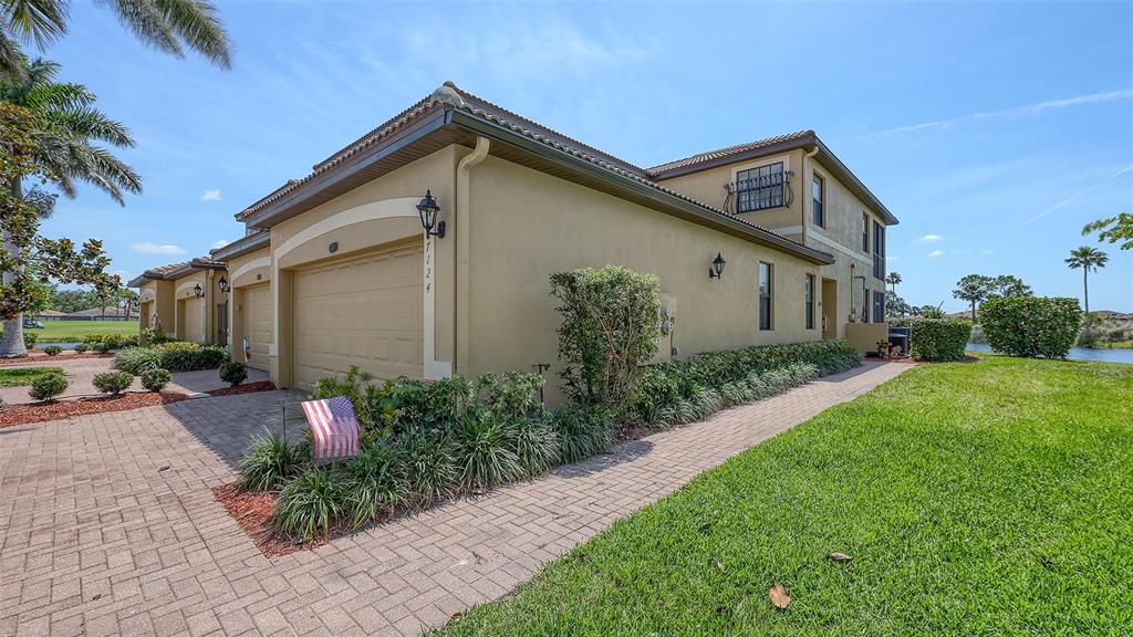 A two-story tan stucco townhouse with a paved driveway and a small American flag — 7124 GRAND ESTUARY TRAIL UNIT 104, BRADENTON, FL