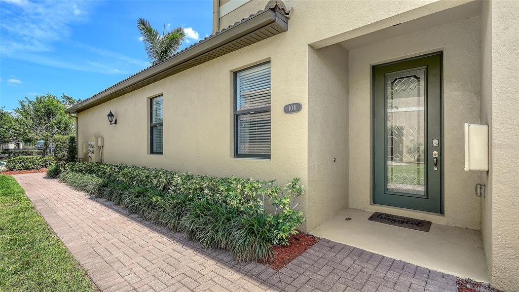 A paved walkway leads to the front entrance of a tan stucco house with landscaping — 7124 GRAND ESTUARY TRAIL UNIT 104, BRADENTON, FL