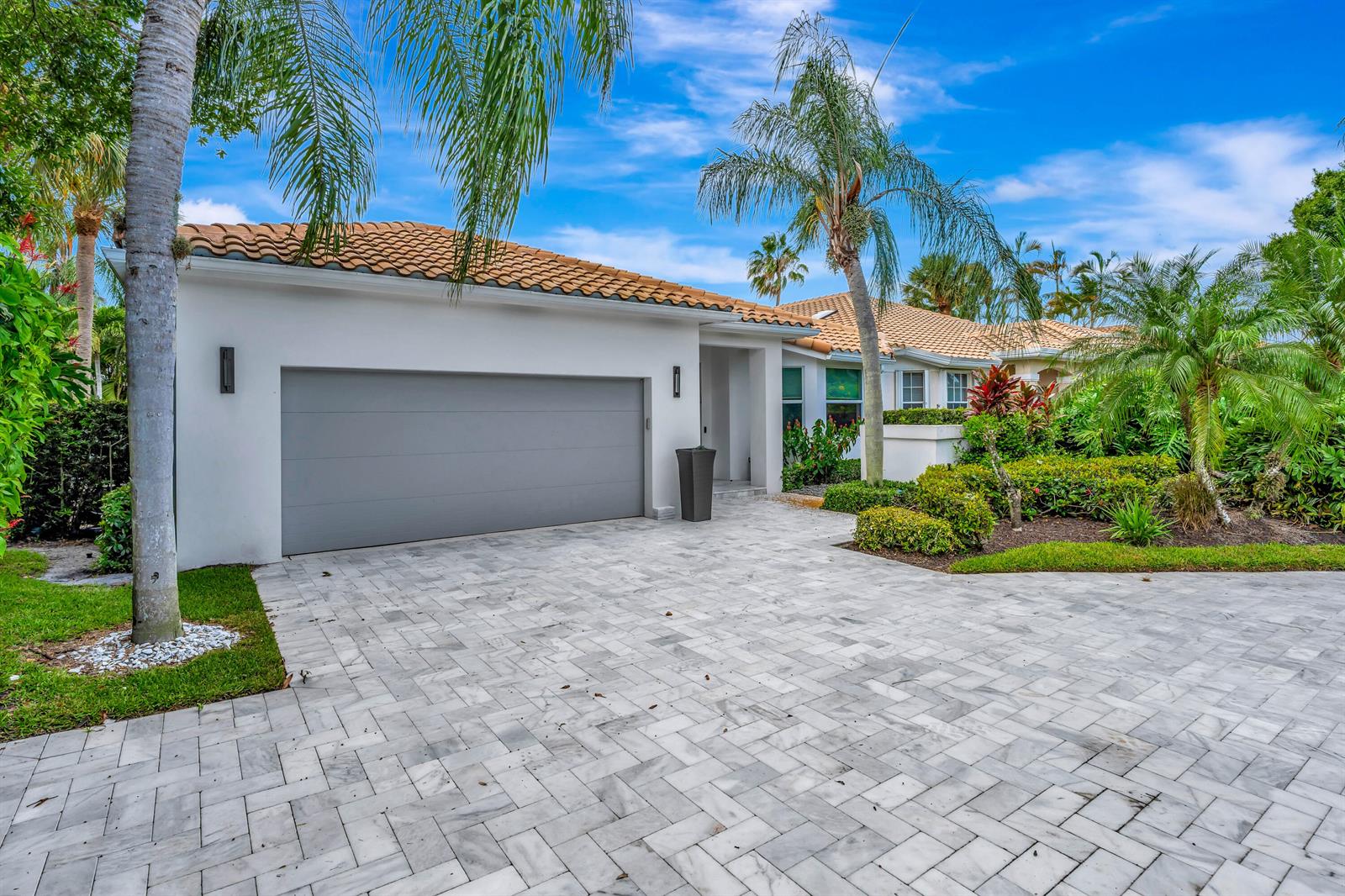 A white house with a gray garage door, tile roof, and paved driveway surrounded by palms — 124 Golf Village Boulevard, Jupiter FL 33458, JUPITER, FL