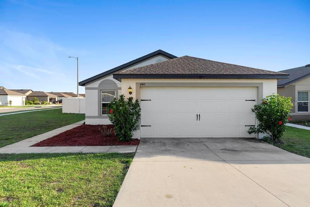 A single-story house exterior features a two-car garage, light-colored stucco walls, and a shingled roof — 440 KENSINGTON VIEW DRIVE, WINTER HAVEN, FL