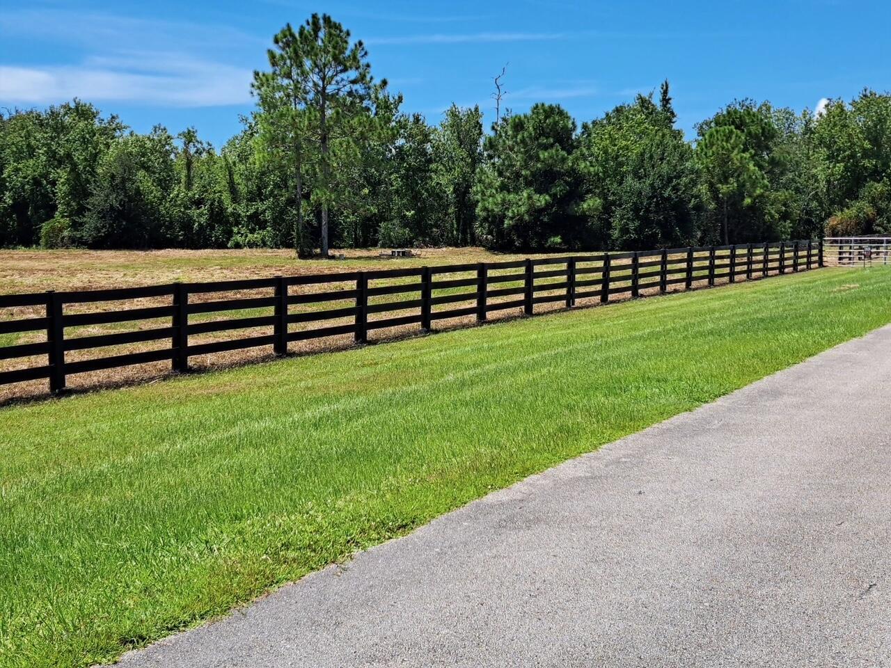A black four-rail wooden fence borders a grassy field next to an asphalt driveway — 13520 SE 22nd Lane, Okeechobee FL 34974, OKEECHOBEE, FL