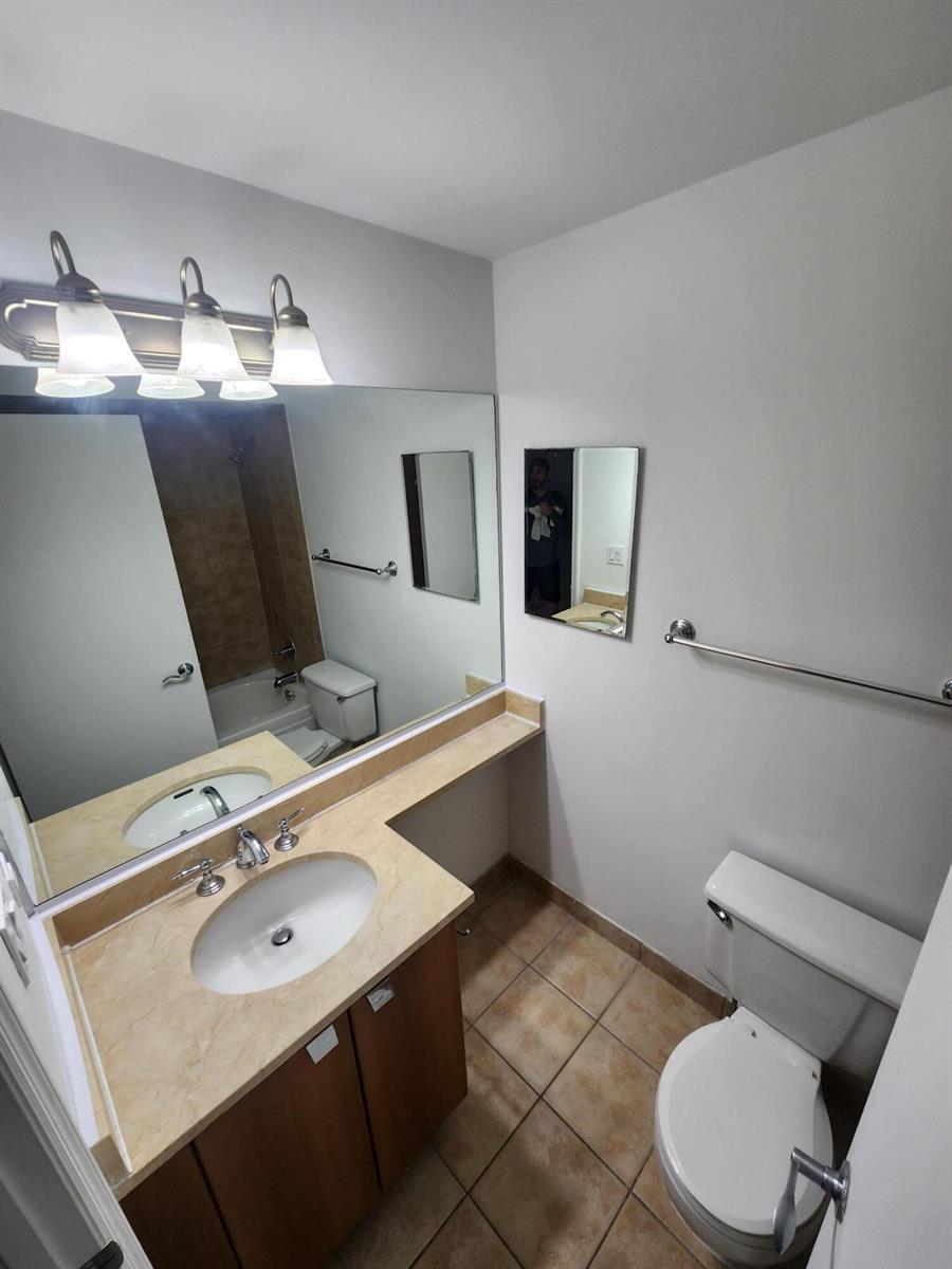 A bathroom features a vanity with a beige countertop, a large mirror, and tiled flooring — 1020 Crystal Way # F, Delray Beach FL 33444, DELRAY BEACH, FL
