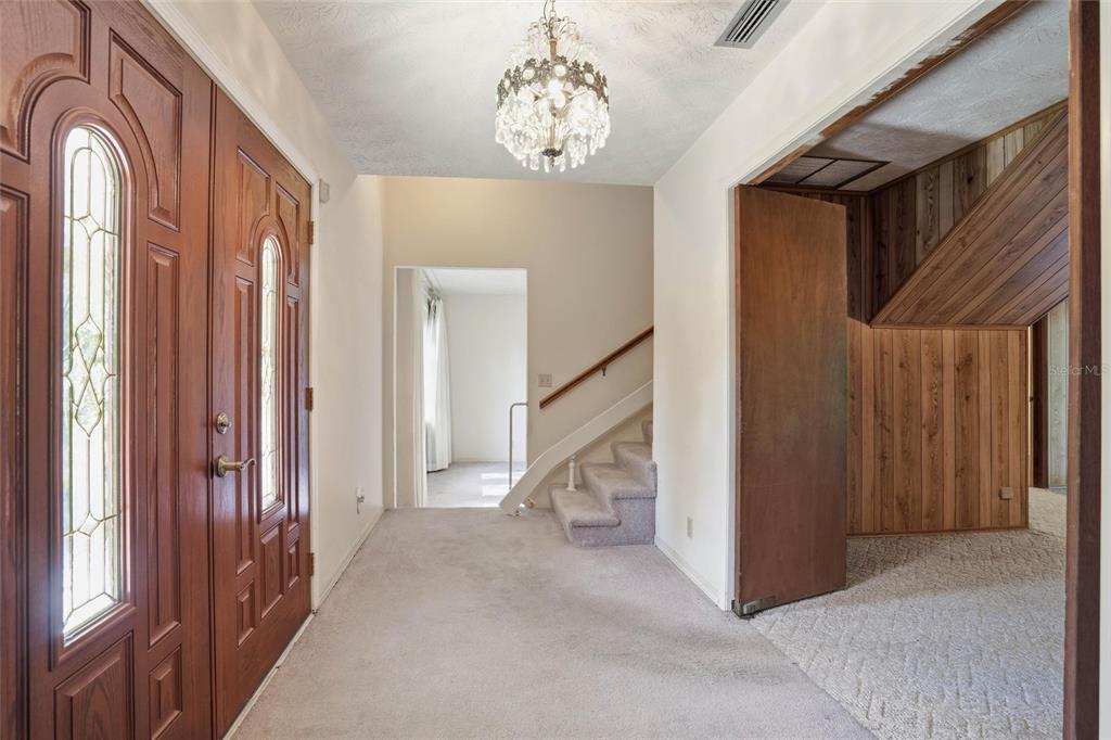 An entryway features a crystal chandelier, carpeted floors, a wooden door, and a staircase — 2406 ELBOW ROAD, ORANGE PARK, FL