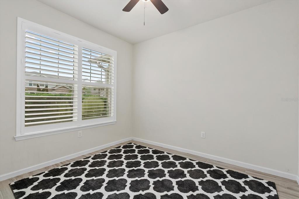 An empty room features light-colored walls, a ceiling fan, plantation shutters, and a patterned rug — 3331 CHESTERTOWN LOOP, BRADENTON, FL