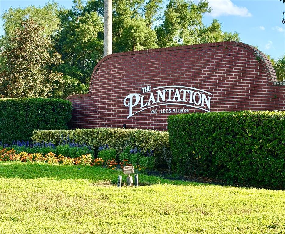 A brick wall displays the sign for The Plantation at Leesburg behind a landscaped garden — 4908 CYPRESS HEAD COURT, LEESBURG, FL