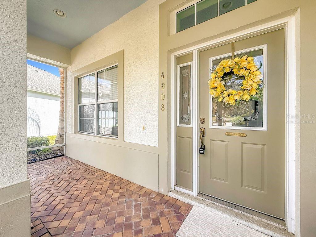 A front porch with brick flooring leads to a tan door with a yellow wreath — 4908 CYPRESS HEAD COURT, LEESBURG, FL