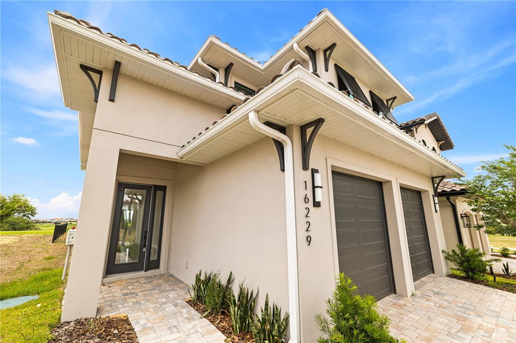 A beige house exterior features a front door, two garage doors, and a paved walkway — 16229 RAVENNA COURT, BELLA COLLINA, FL