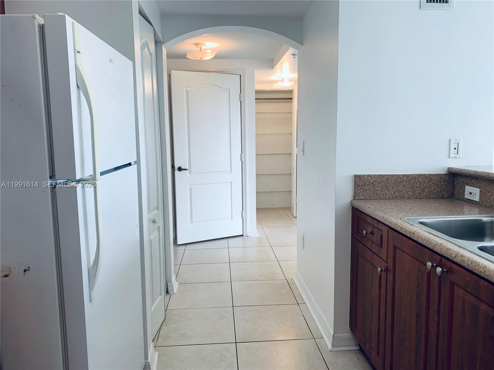 A kitchen with a white refrigerator and brown cabinets leads to a hallway with shelving — 406 NW 22nd Ave # 702, Miami FL 33125, MIAMI, FL