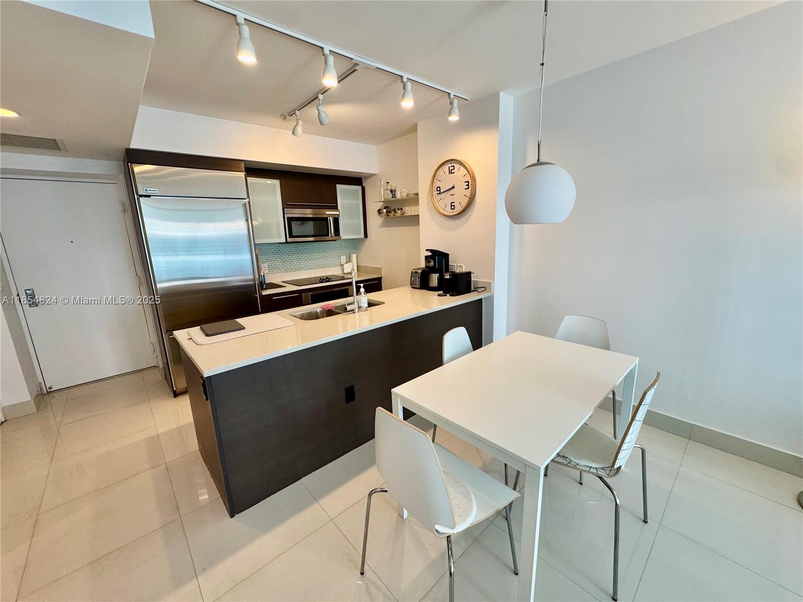 A modern kitchen features dark cabinetry, stainless steel appliances, a white island, and dining area — 500 Brickell Ave # 1204, Miami FL 33131, MIAMI, FL