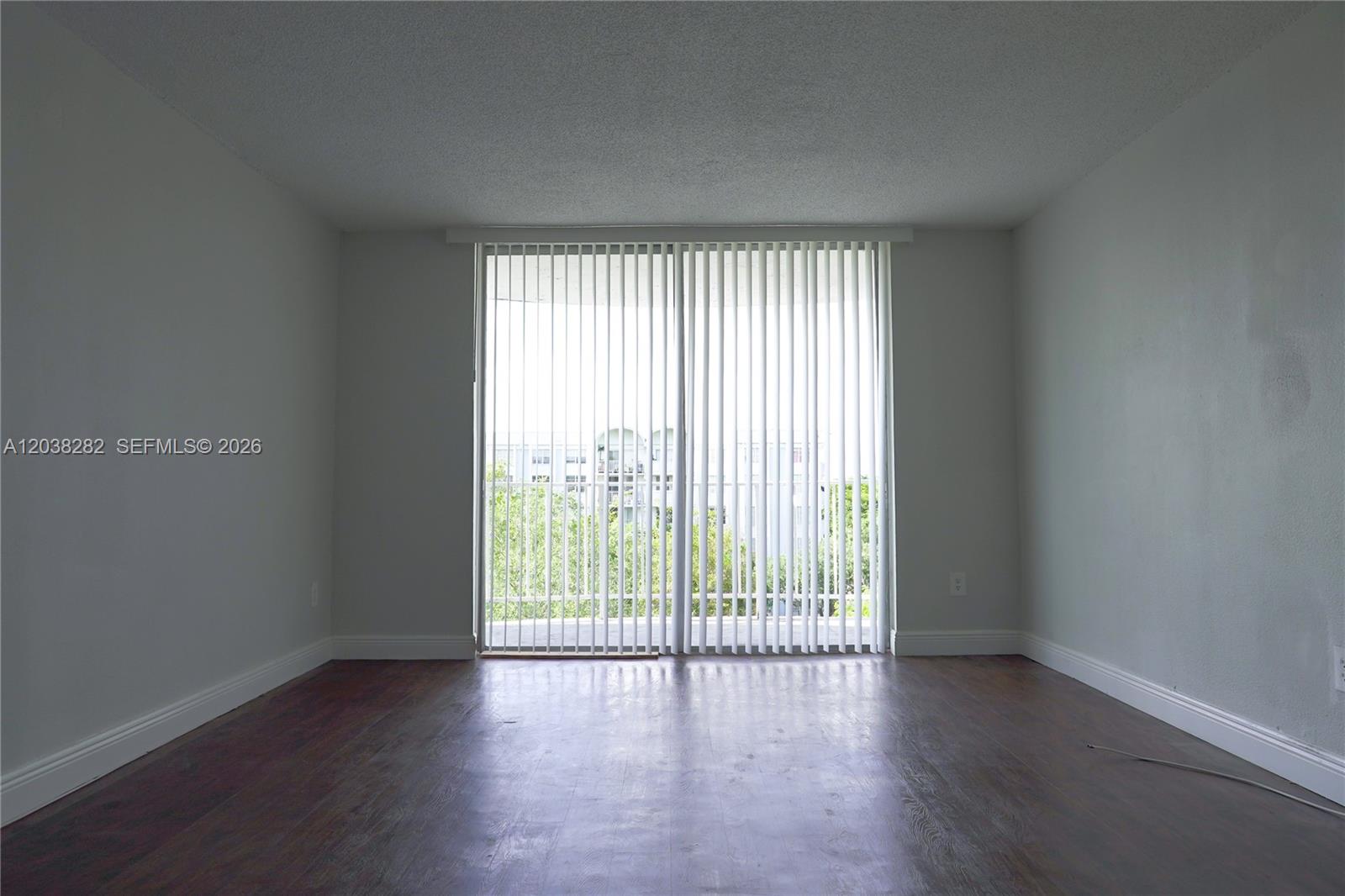 An empty room features dark wood flooring, light gray walls, and vertical blinds over sliding doors — 482 NW 165th St Rd # A504, Miami FL 33169, MIAMI, FL