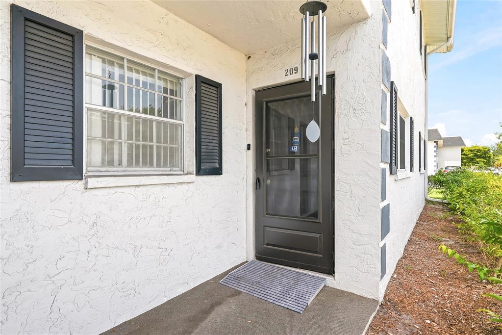 A white stucco building exterior features a dark front door, shutters, and a hanging chime — 202 46TH AVENUE TERRACE W UNIT #209, BRADENTON, FL