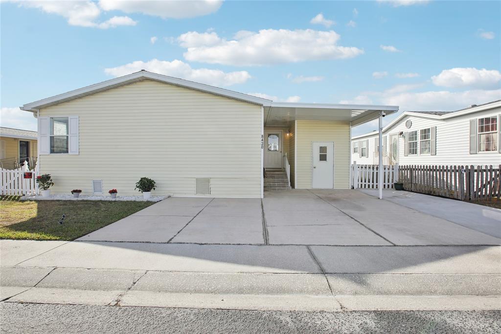 A light yellow manufactured home features a concrete driveway, a carport, and a white fence — 8420 FANTASIA PARK WAY, RIVERVIEW, FL