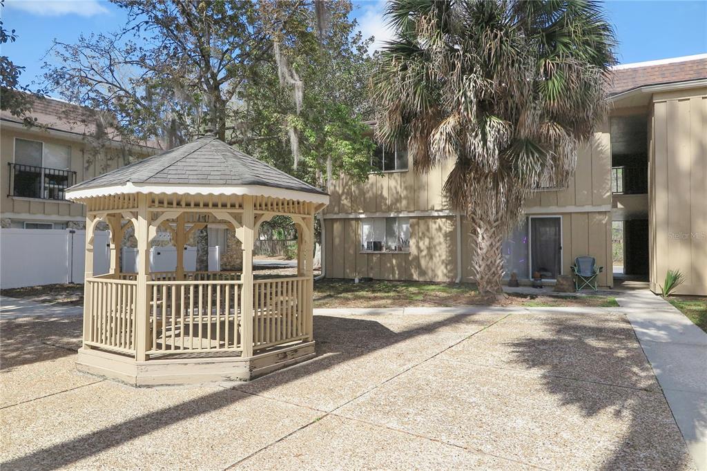 A beige wooden gazebo sits on a concrete patio in front of a two-story apartment building — 507 NW 39TH DRIVE UNIT 210, GAINESVILLE, FL