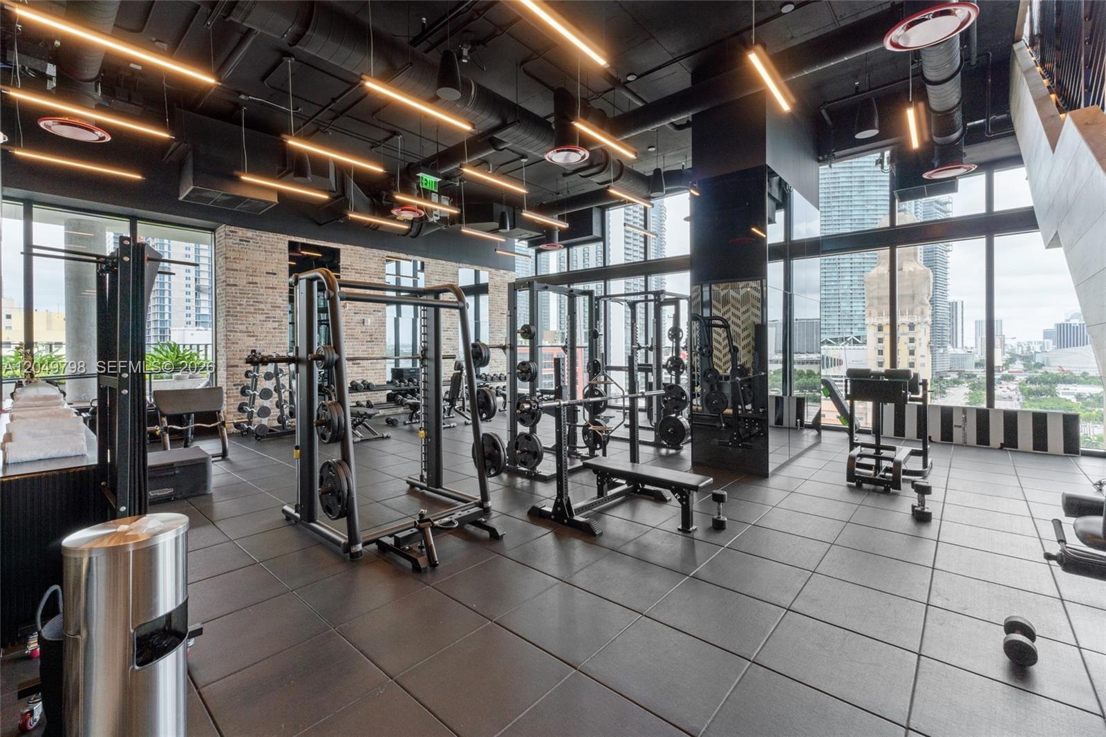A modern gym features weight racks, black tile flooring, exposed ceiling ducts, and floor-to-ceiling windows — 398 NE 5th St # 4719, Miami FL 33132, MIAMI, FL