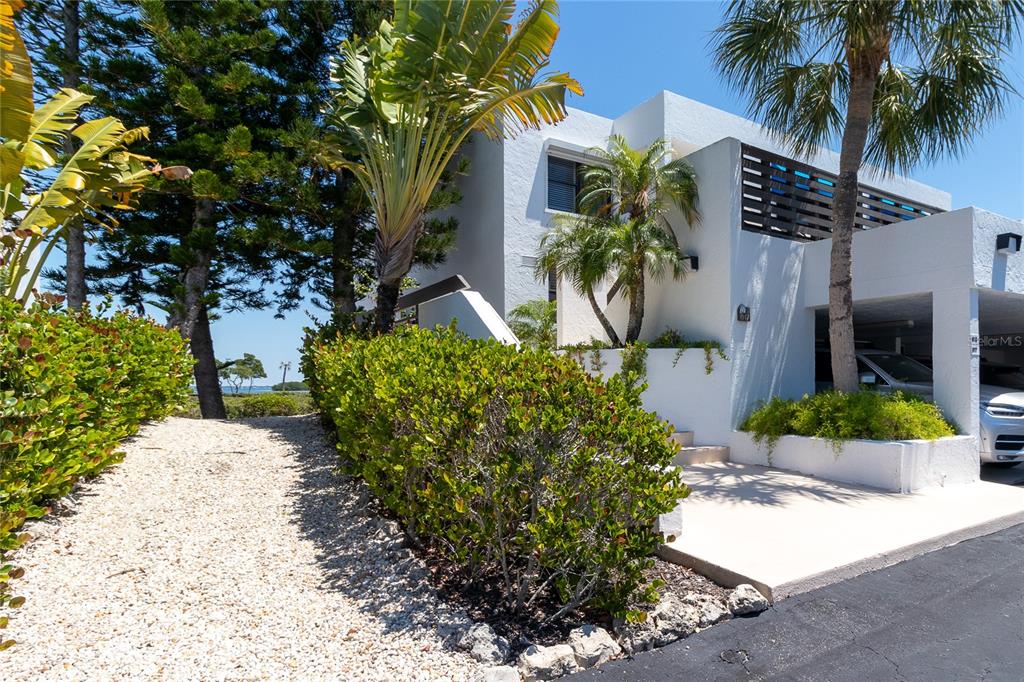 A white multi-story house features a gravel path, lush landscaping, and a covered parking area — 817 BAYPORT WAY UNIT 817, LONGBOAT KEY, FL