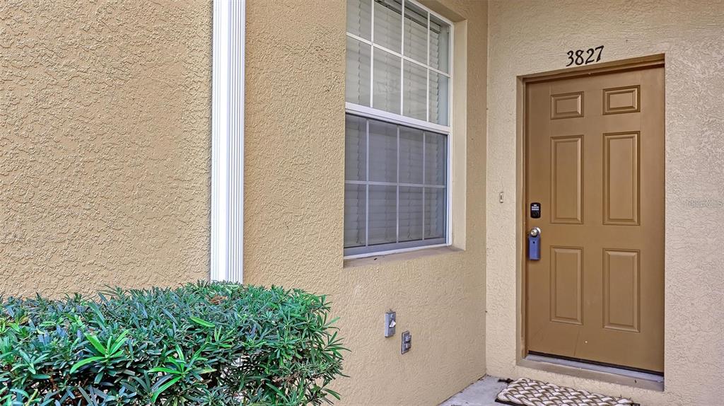 A tan exterior wall features a brown front door, a window, and a green bush — 3827 PARKRIDGE CIRCLE UNIT 1-102, SARASOTA, FL